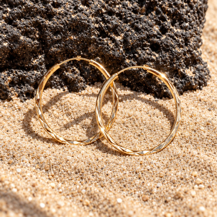 Geometrische Creolen in Champagner-Gold liegend im Sand vor dunklem Vulkanstein