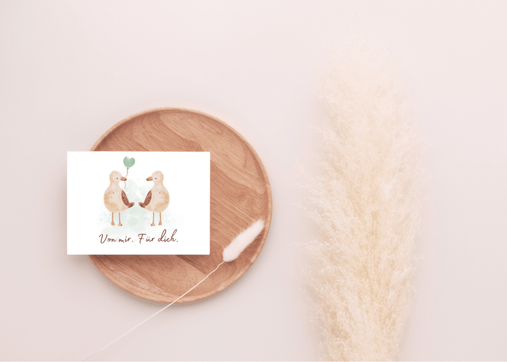 Carte cadeau motif oiseau marin posée sur un plateau rond en bois avec une fleur séchée