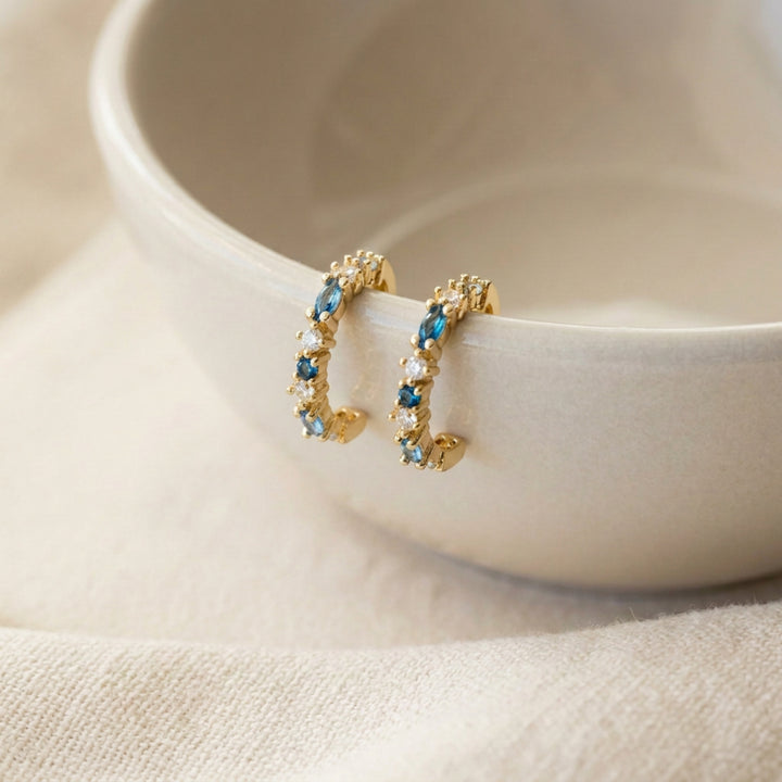 Gold Blue Crystal huggie hoop earrings hanging on the rim of a beige ceramic dish