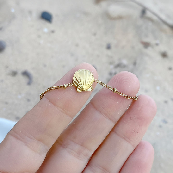 Goldenes Armband mit Muschel, Jakobsmuschel Anhänger Charm aus hautverträglichem wasserdichtem Edelstahl auf Hand vor Sand