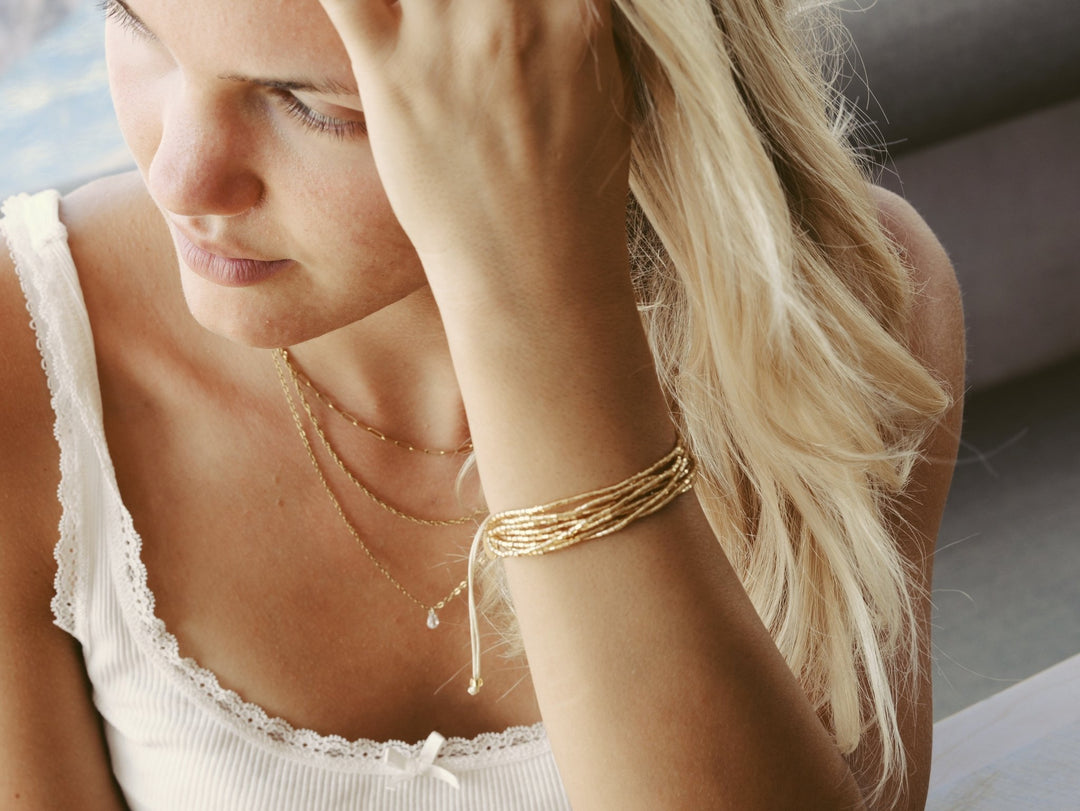 Goldener Schmuck an schöner junger Frau mit blonden Haaren und weißen Spitzen-Oberteil, Armband und Kette von Alicia&